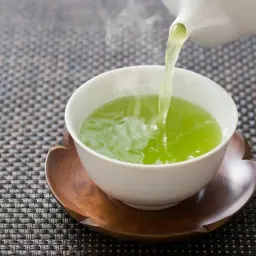 Zielona herbata nalewana z czajniczka do białej filiżanki na drewnianej podstawce.